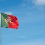 Bandeira de Portugal a ondear num mastro sob um céu azul com nuvens, simbolizando o lançamento do portal Portugal EuroBrief.